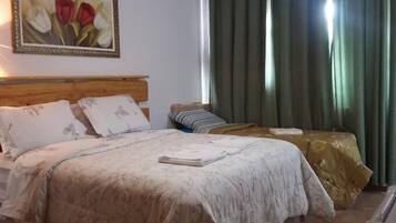Quarto com cabeceira de madeira, duas camas com roupa de cama floral, mesa de cabeceira, mesa lateral, quadro decorativo com flores e cortinas verdes.