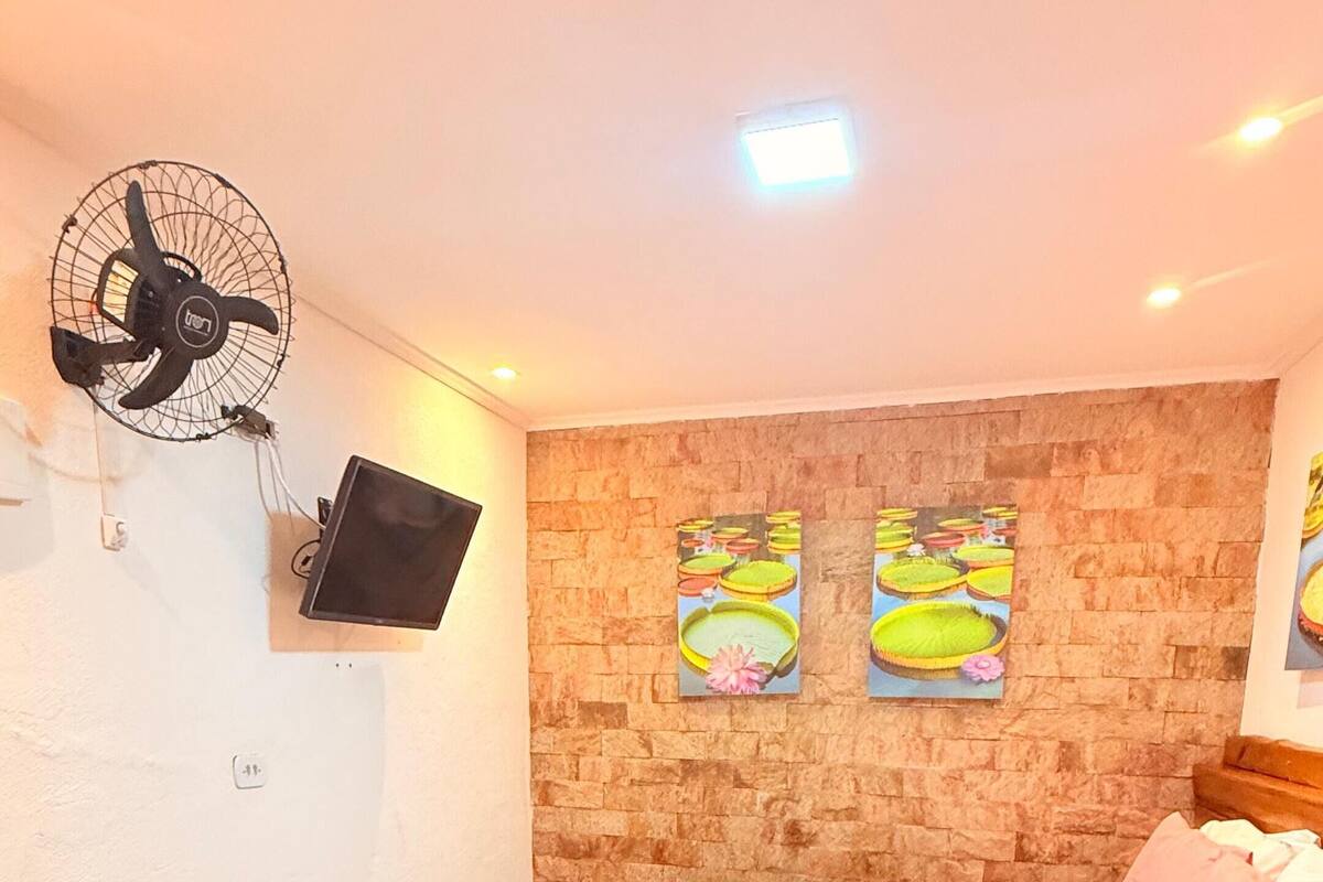 A bedroom with a bed, a wall-mounted fan, a flat-screen TV, and two wall art pieces.