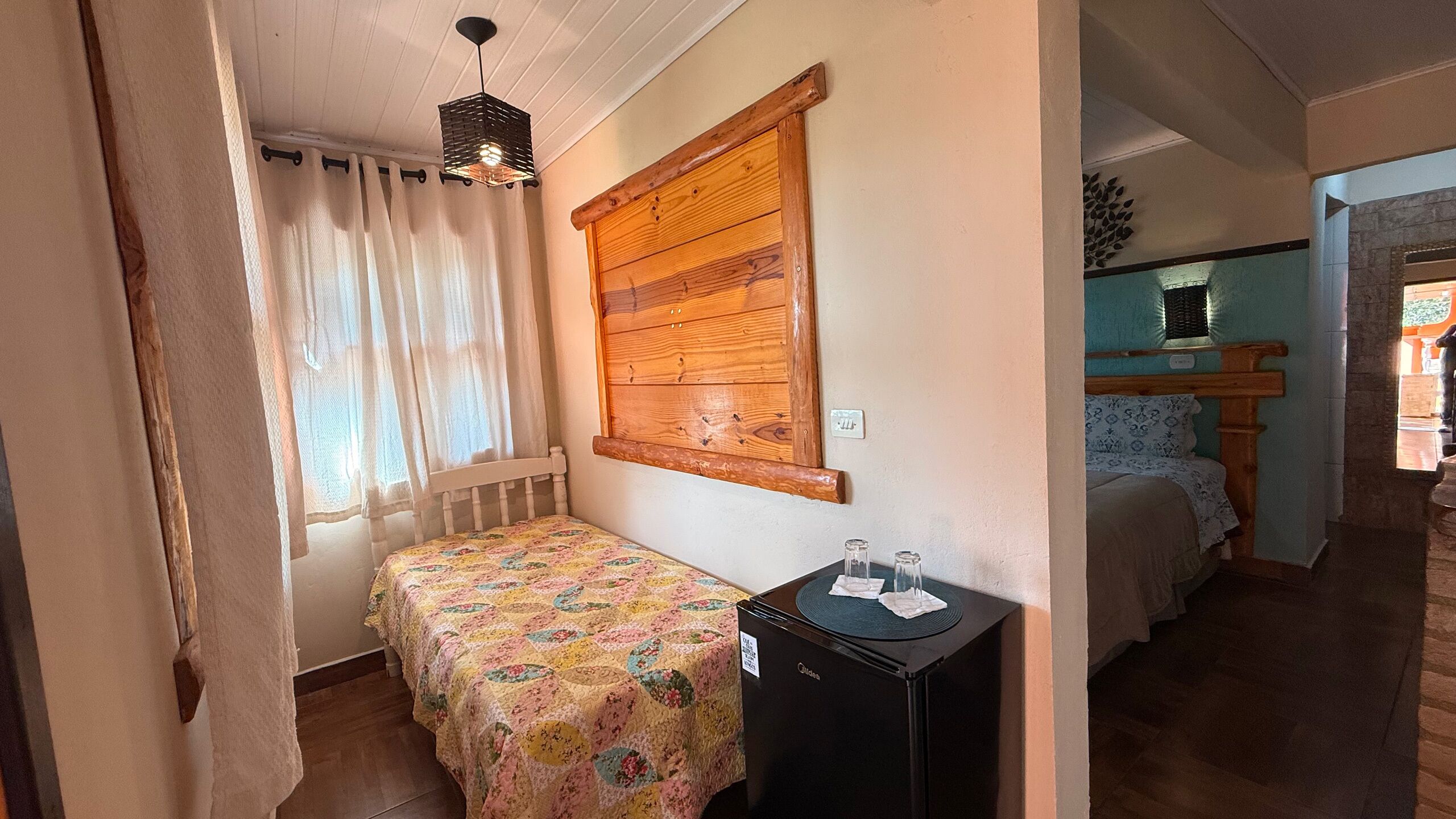 Un dormitorio con cabecera de madera, cama con colcha floral, una mesita de noche con lámpara y una ventana con cortinas translúcidas.
