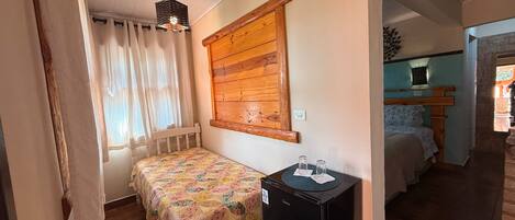 Un dormitorio con cabecera de madera, cama con colcha floral, una mesita de noche con lámpara y una ventana con cortinas translúcidas.