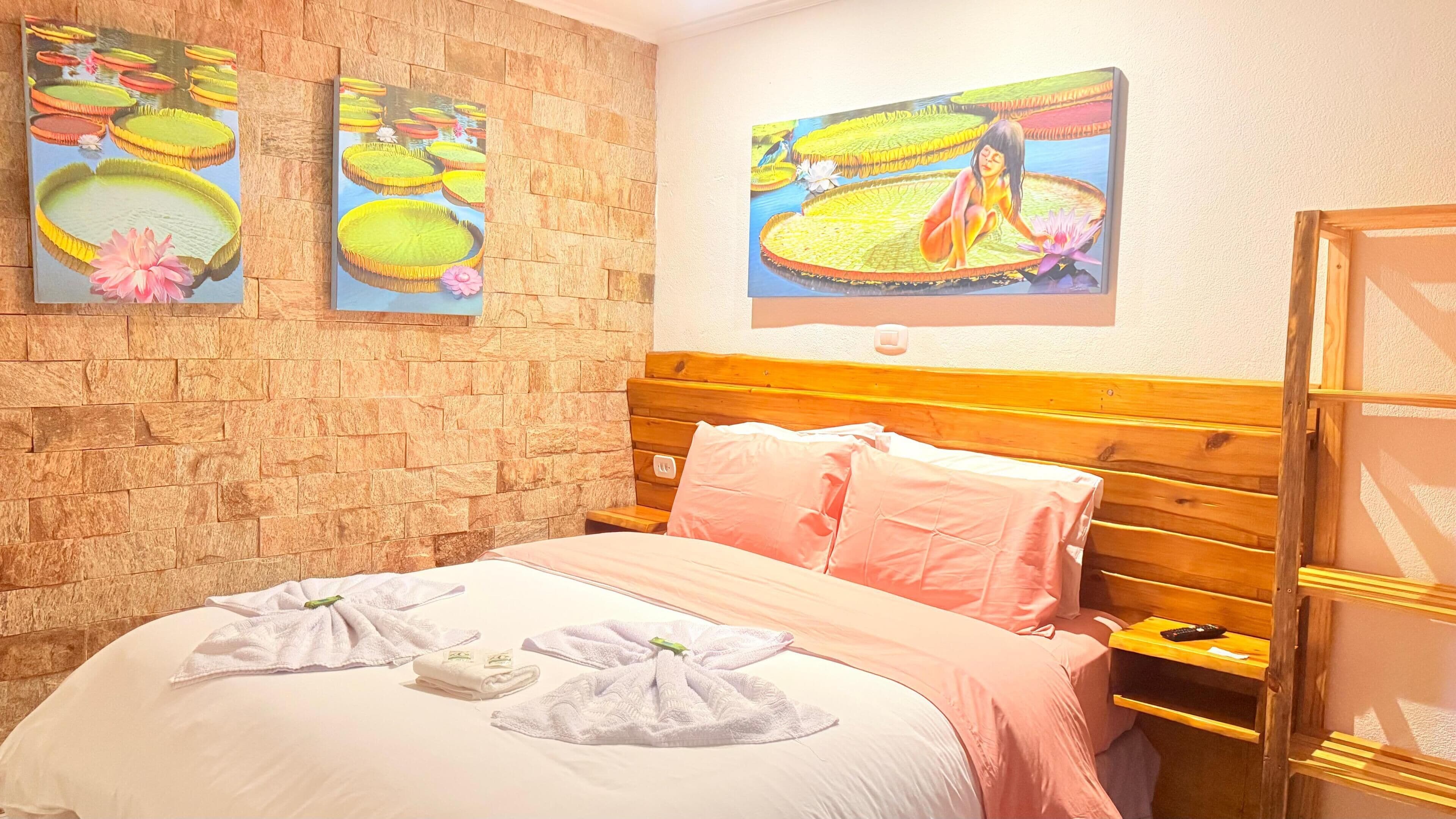 Quarto de hotel com parede de pedra, cama de madeira com roupa de cama rosa e obra de arte retratando nenúfares.
