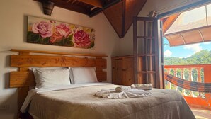 Ein Schlafzimmer mit hölzernem Kopfteil, einem großen floralen Gemälde, Blick auf eine üppige Landschaft durch eine offene Tür und einer Hängematte auf dem Balkon.