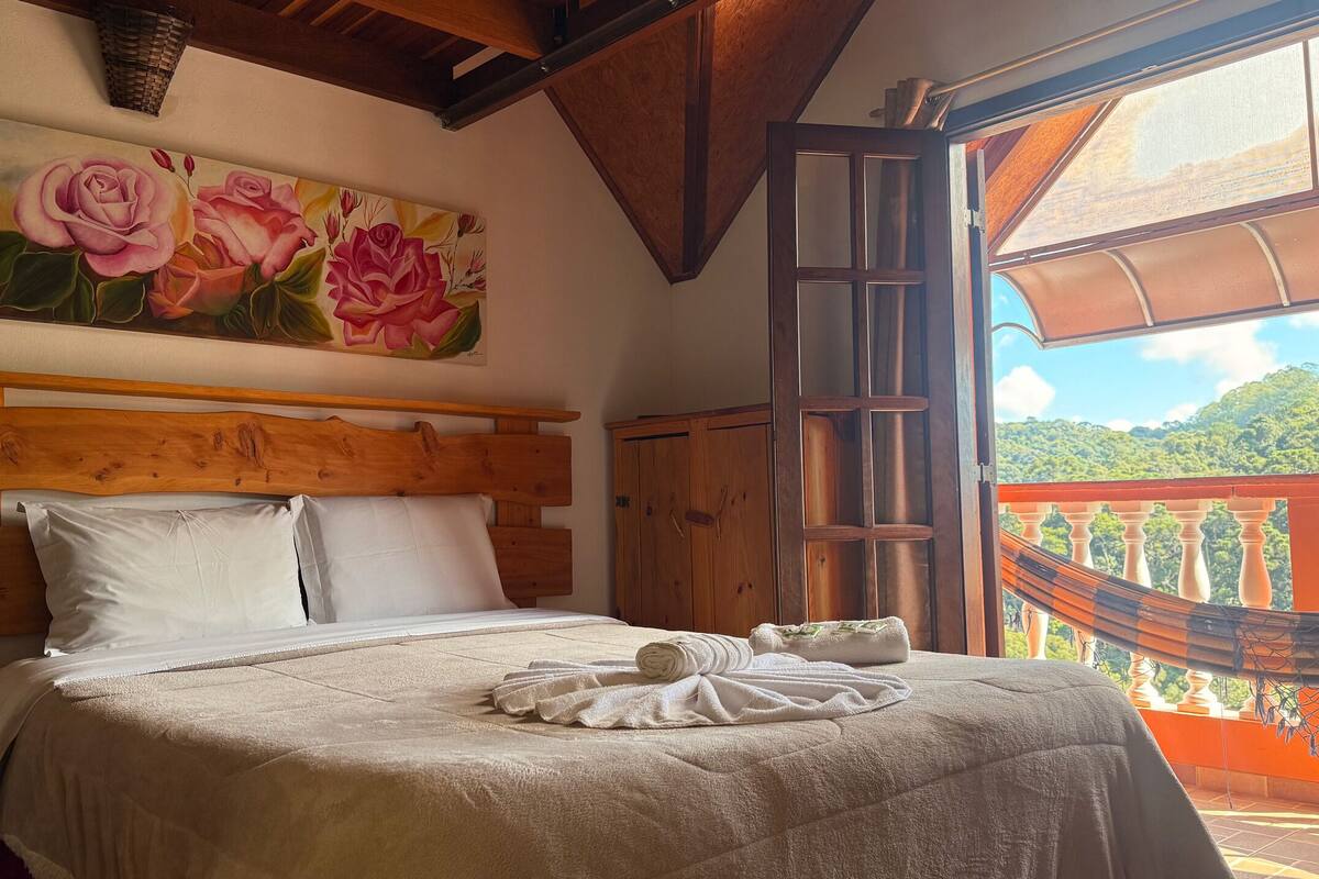 A bedroom with a wooden headboard, a large floral painting, a view of a lush landscape through an open door, and a hammock on the balcony.