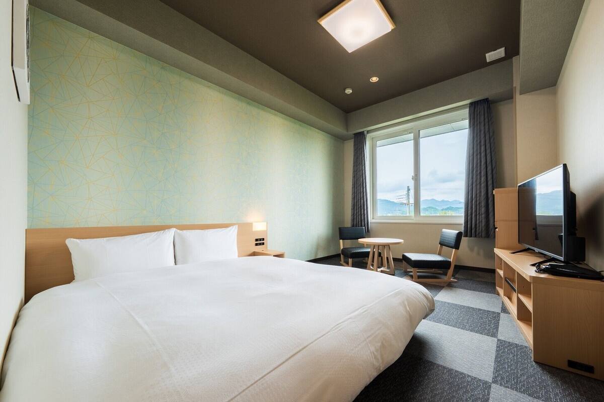 A hotel room with a large bed, a desk with a television, a chair, and a view of the mountains through the window.