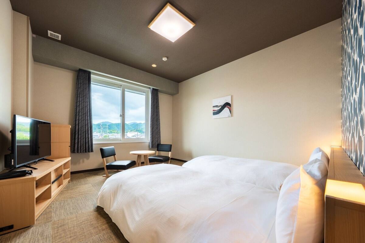 A hotel room with a large bed, a desk with a TV, two chairs, a small table, and a window with a view of the mountains.