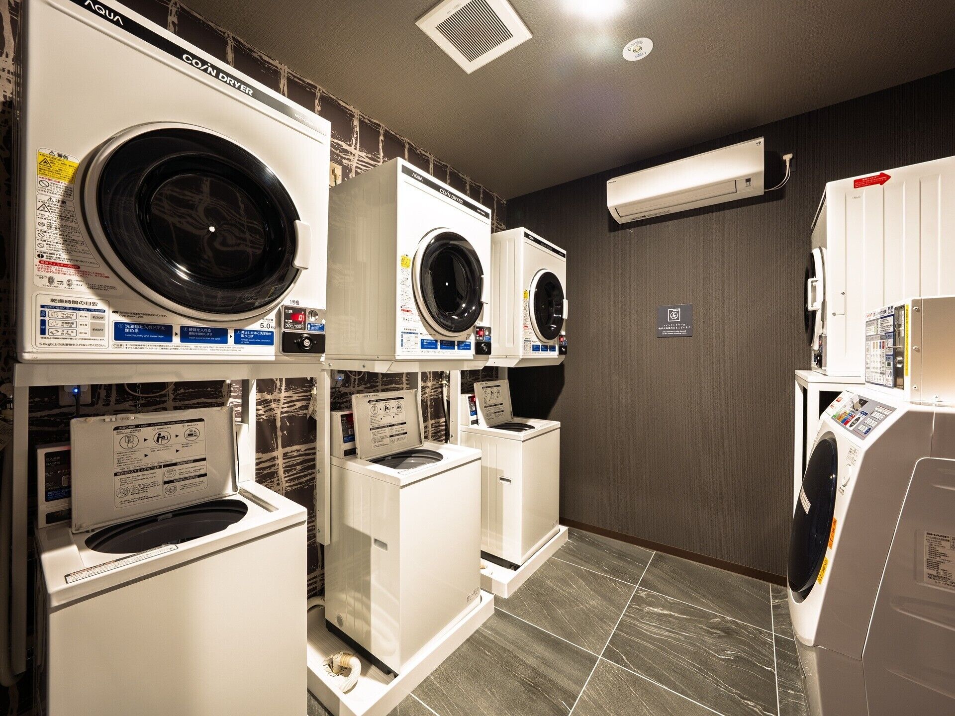 A laundry facility with multiple washing machines and dryers, a tiled floor, and a wall-mounted air conditioning unit.