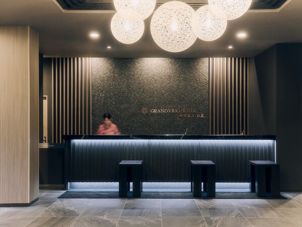Zona de recepción de un hotel con un mostrador oscuro, tres taburetes negros y una pared decorativa con el nombre del hotel.