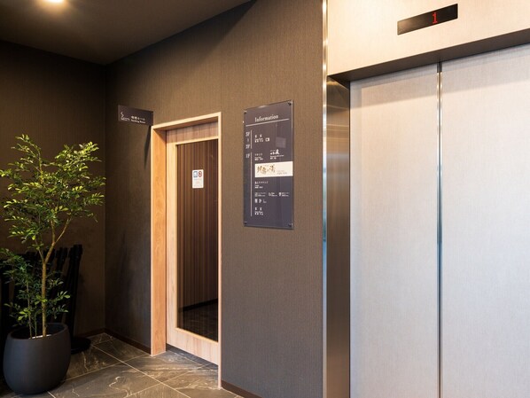 Un ascensor con pantalla digital, un cartel con información de habitaciones y una planta en maceta.