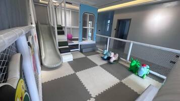 Un espace de jeux pour enfants avec un toboggan, des tapis de jeu souples et une zone de jeux délimitée par un filet.