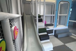 Zona de juegos infantiles con tobogán, estructuras de juego cubiertas con red y escalones acolchados.