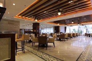 A spacious hotel lobby with a modern design, featuring a central seating area with tables and chairs, a patterned floor, and a ceiling with linear lighting.
