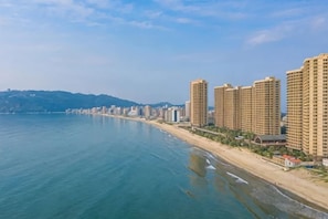 A coastal cityscape with high-rise buildings along a beach, a clear sky, and distant hills. - Honghai Bay Haihang Holiday Hotel (Huidong)