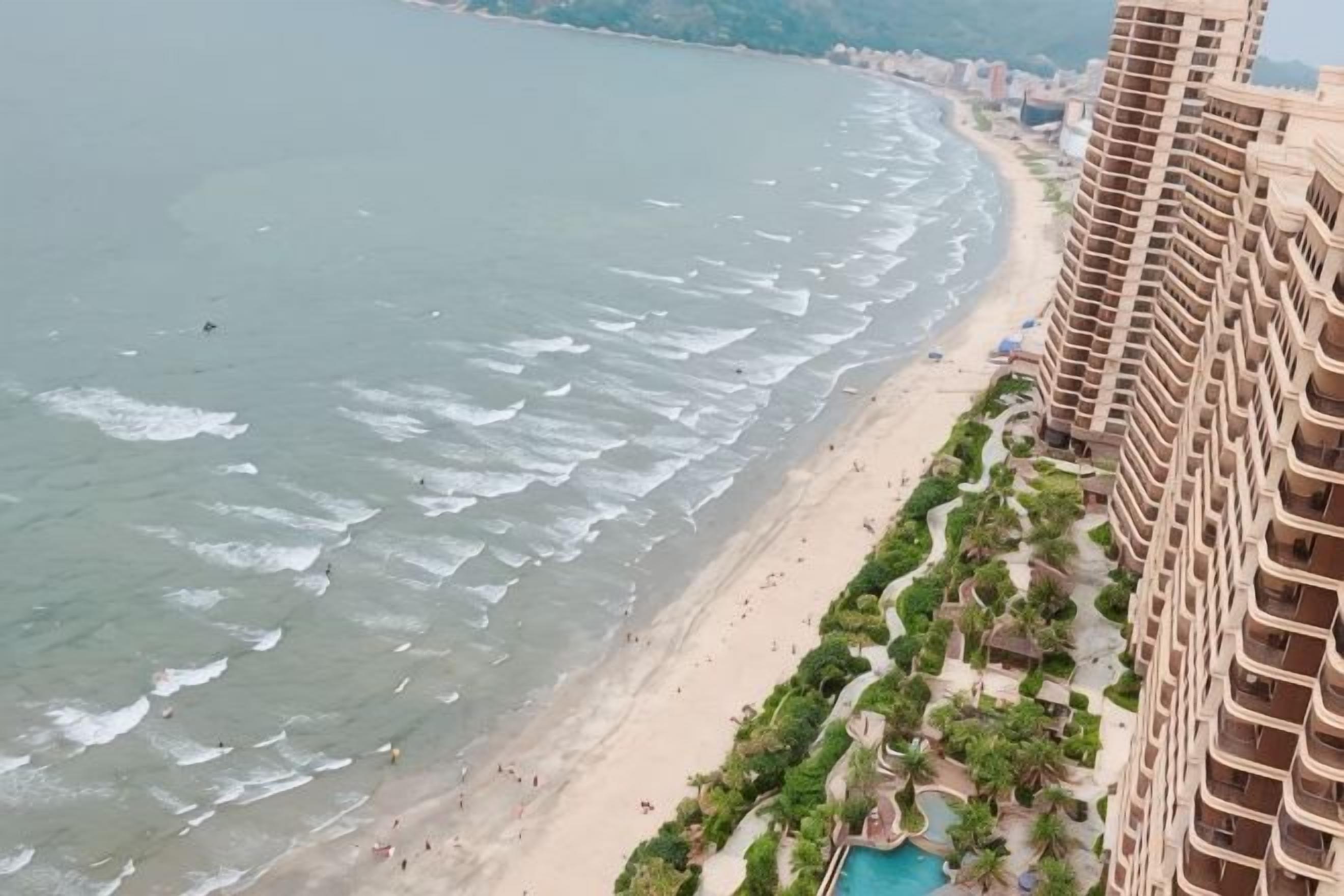 A coastal hotel with a beachfront view, a pool, and a promenade with greenery.