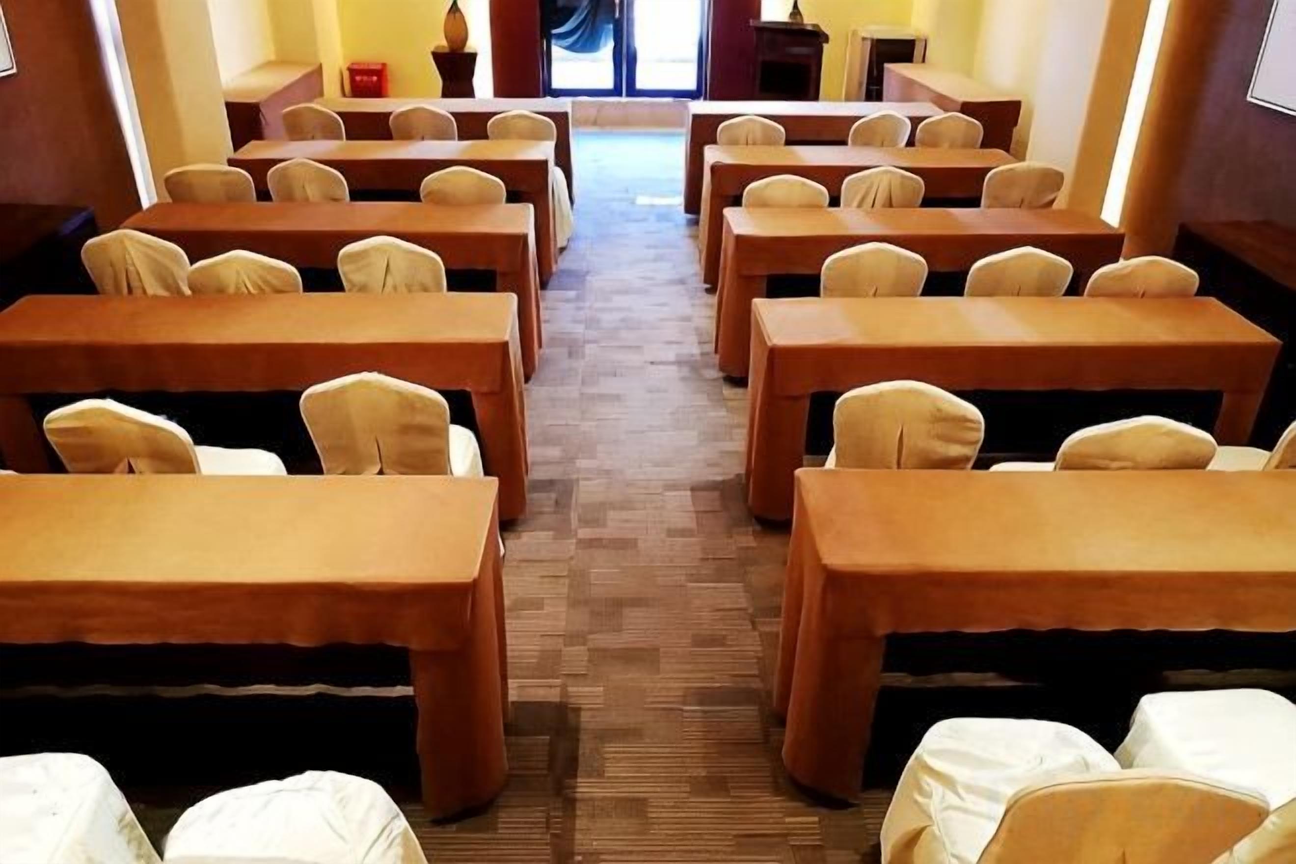 Un salón de conferencias con filas de mesas y sillas, piso alfombrado y vista al exterior a través de una puerta.
