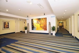 Un pasillo de hotel con alfombra a rayas, un cuadro abstracto grande y plantas en macetas.