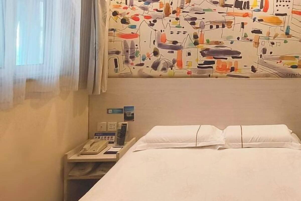 Ein Hotelzimmer mit einem Bett, einem Nachttisch mit Telefon und Lampe sowie einer Wand mit einem farbenfrohen, abstrakten Wandbild.