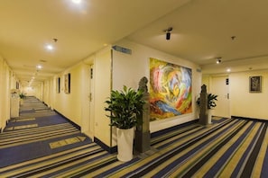 En hotelgang med tæppe, et stort farverigt billede på væggen, potteplanter og et skilt, der viser vejen til lobbyen.