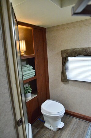 A compact bathroom with a toilet, a wooden cabinet with folded towels, and a window with blinds. - Casa Coqui Camper, with WiFi, & AC, 3 miles from Falls Creek Falls State Park (Pikeville)