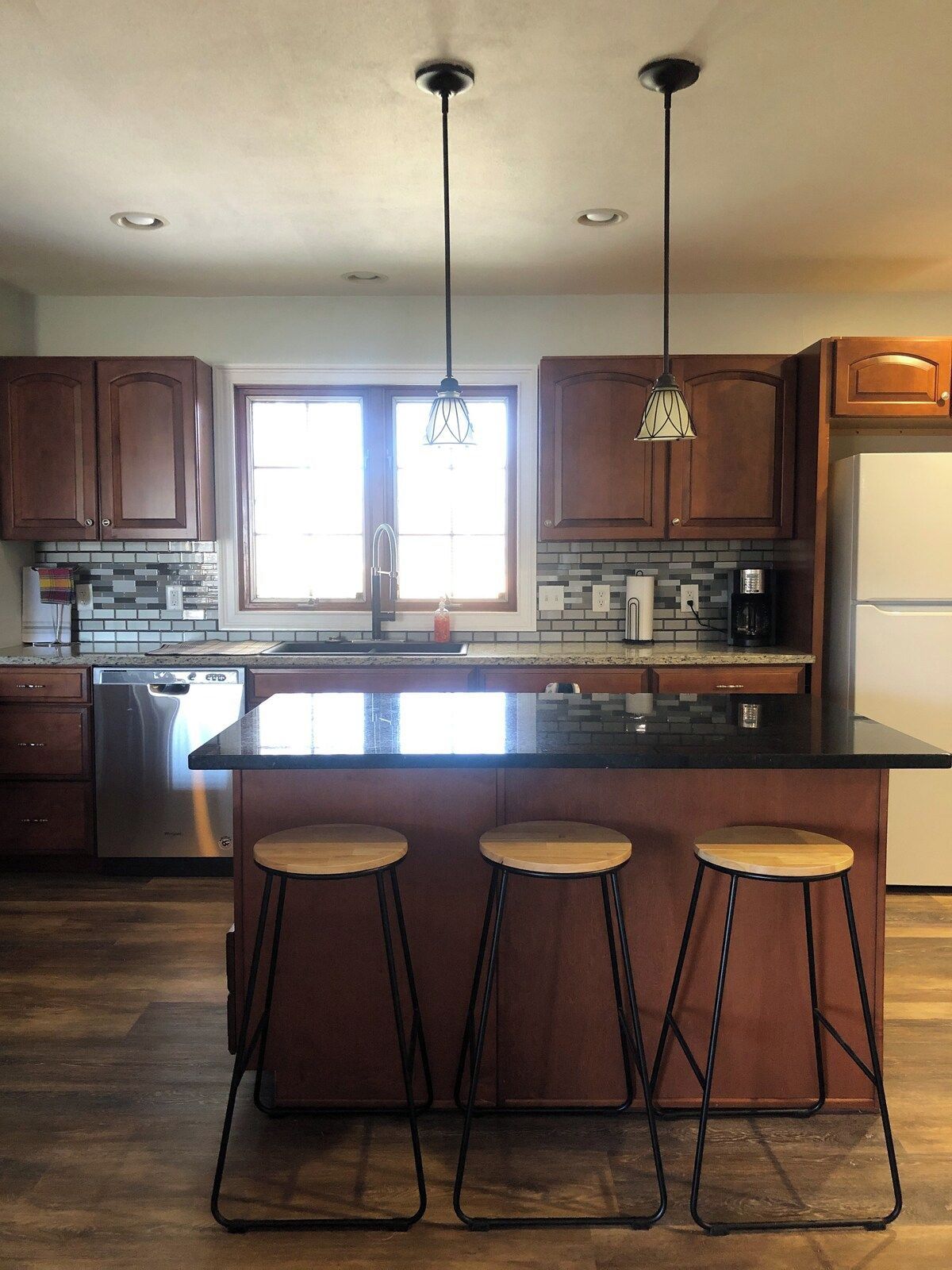 A modern kitchen with wooden cabinets, a stainless steel dishwasher, a large island with stools, pendant lighting, and a window providing natural light.