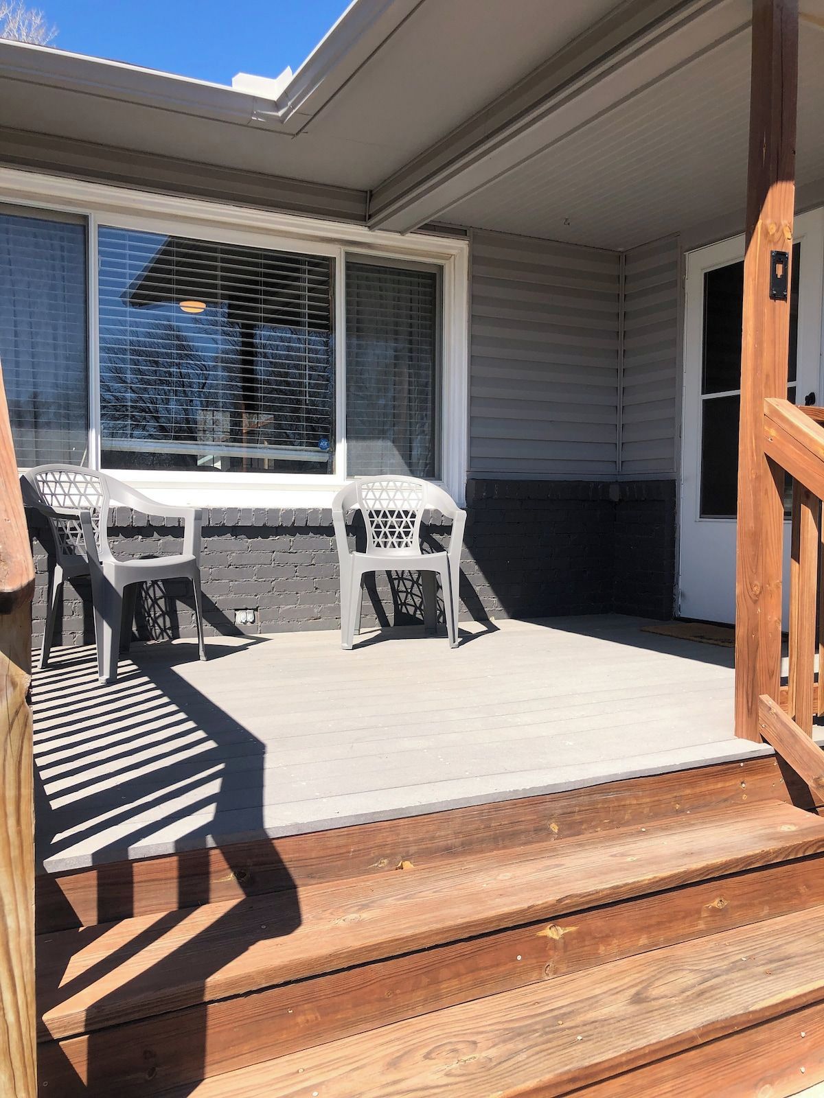 A covered outdoor area with a wooden deck, a table with chairs, and a window with blinds.