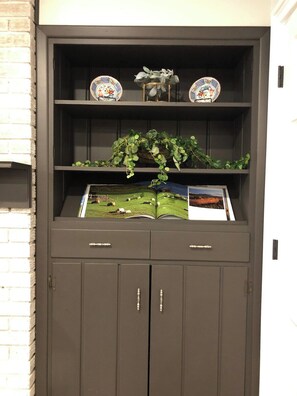 A dark-colored cabinet with two open shelves, one holding decorative plates and the other with a basket of green plants. Below, there is a closed cabinet with two drawers.