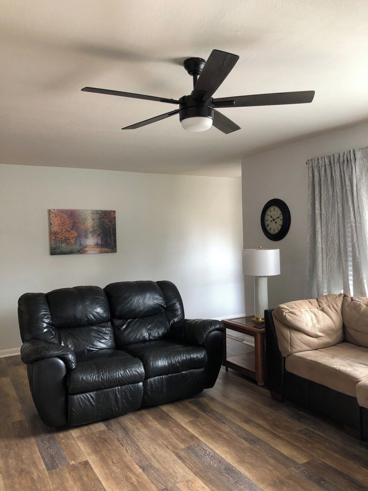 A living room with a black leather sofa, a beige sofa, a wooden floor, a ceiling fan, a wall clock, and a painting.