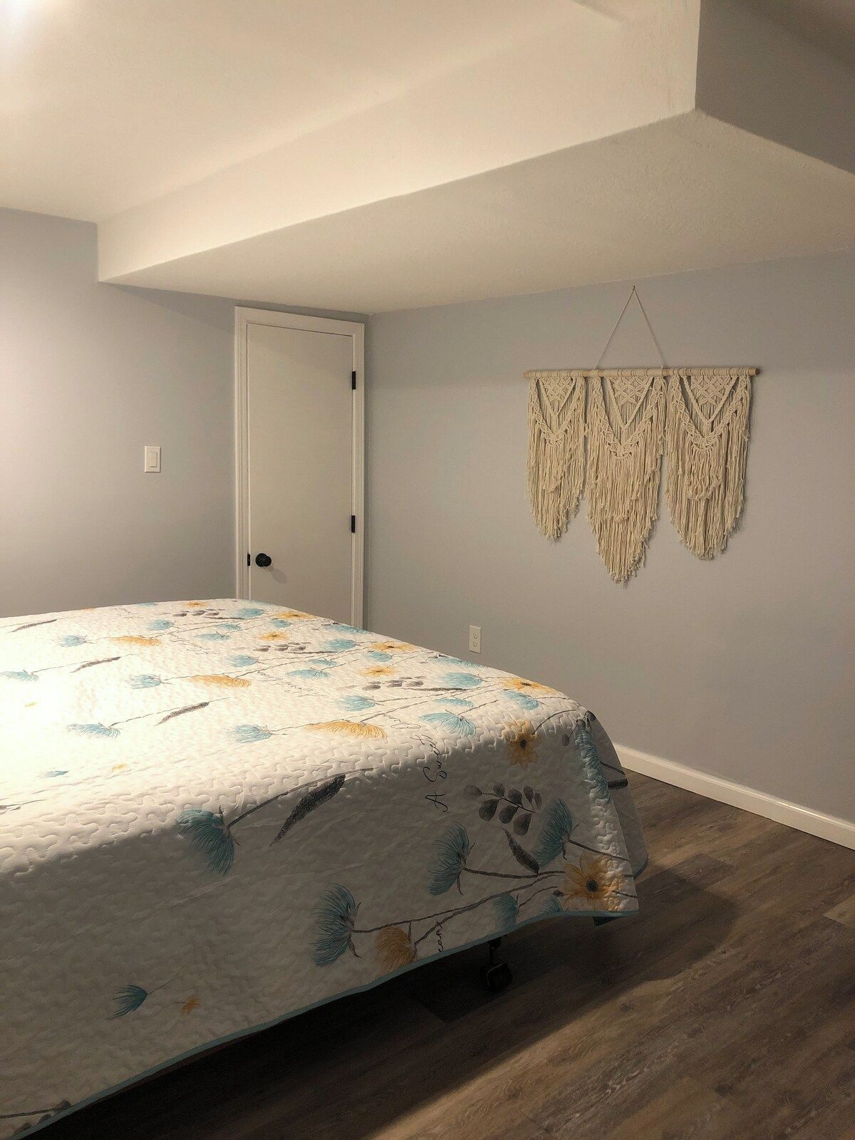 A bedroom with a bed, a door, and a macrame wall hanging.
