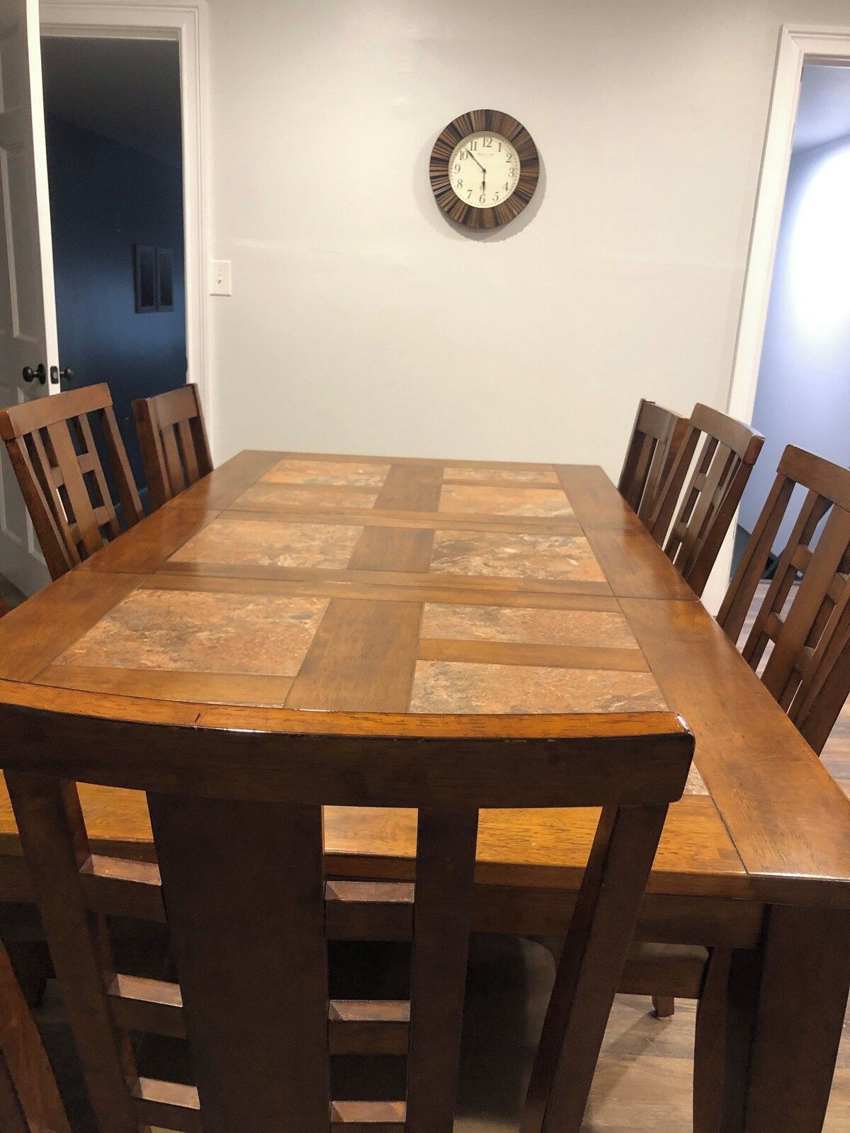 Comedor con una mesa de madera rectangular rodeada por seis sillas de madera. Un reloj de pared es visible sobre la mesa.