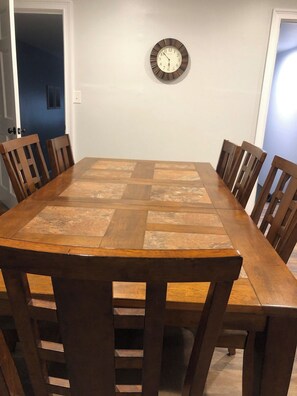 A dining room with a rectangular wooden table surrounded by six wooden chairs. A wall-mounted clock is visible above the table. - Park Place Your Comfortable Stay (Hutchinson)