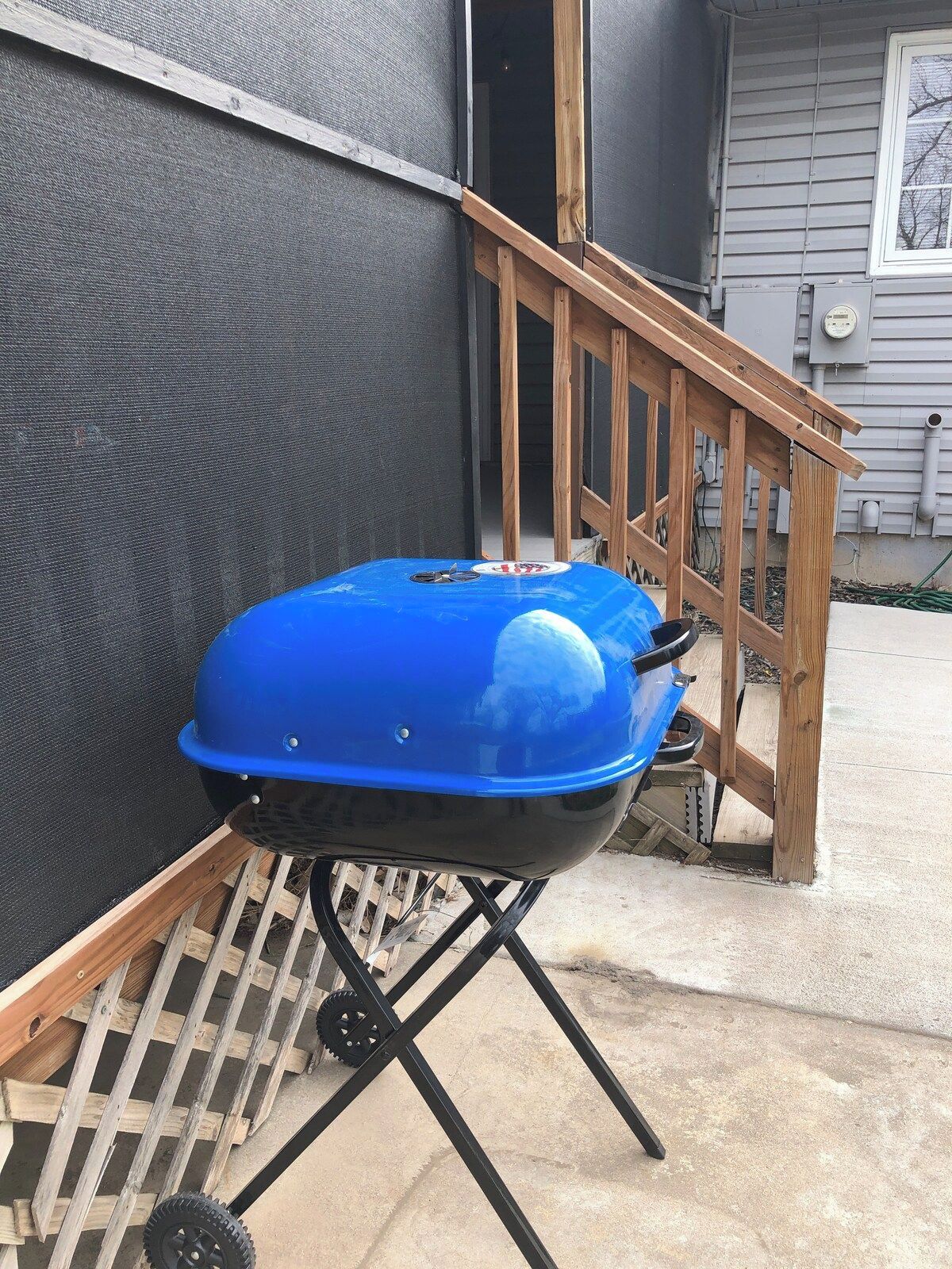 Una parrilla azul portátil con ruedas, colocada sobre un soporte de madera al aire libre.