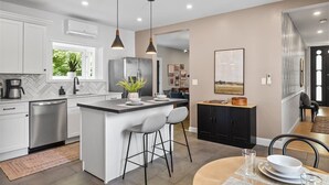 Une cuisine moderne avec des meubles blancs, un îlot central doté de tabourets, un éclairage suspendu et un coin repas avec une table ronde et des chaises.