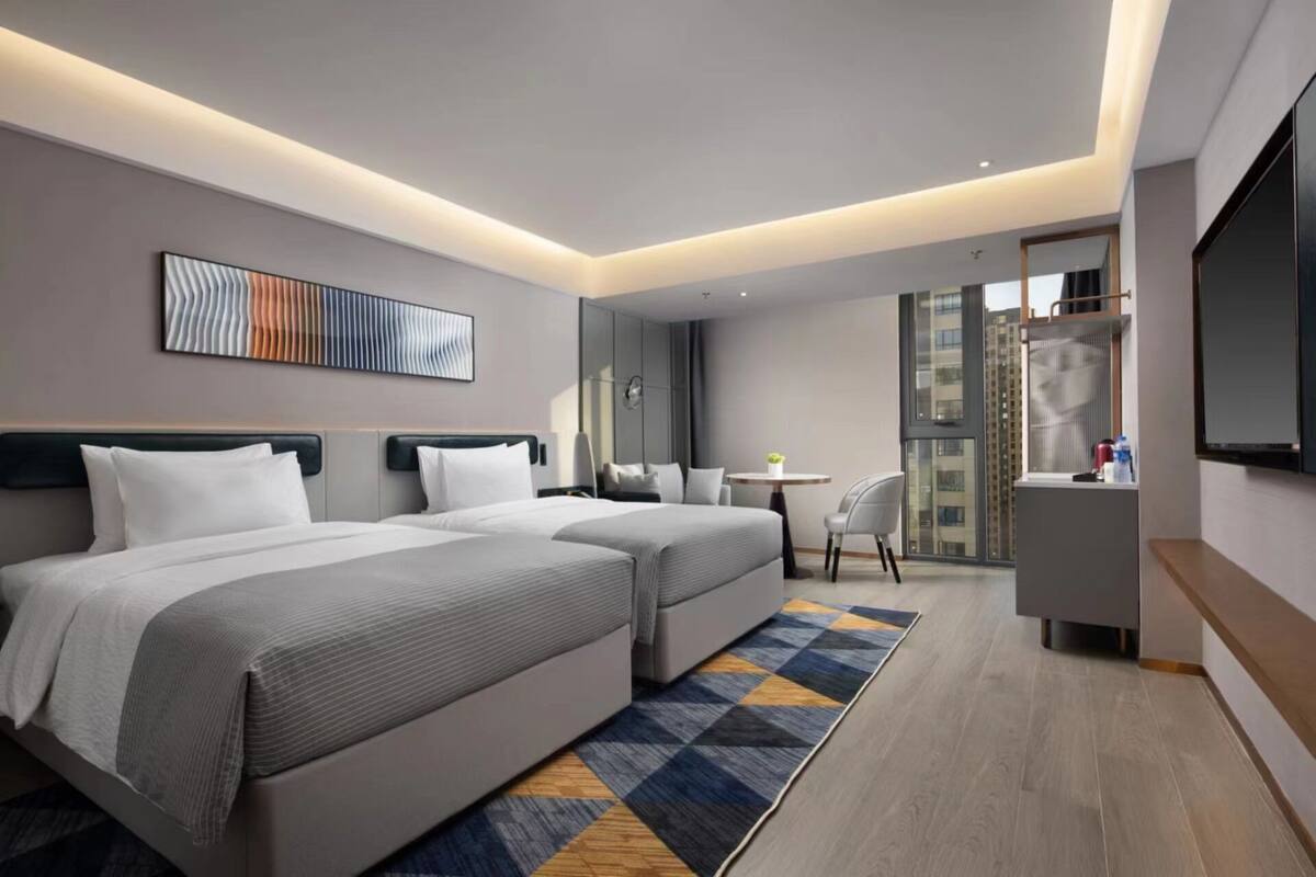 A modern hotel room with two beds, a small table, a chair, a TV, and a view of the city.