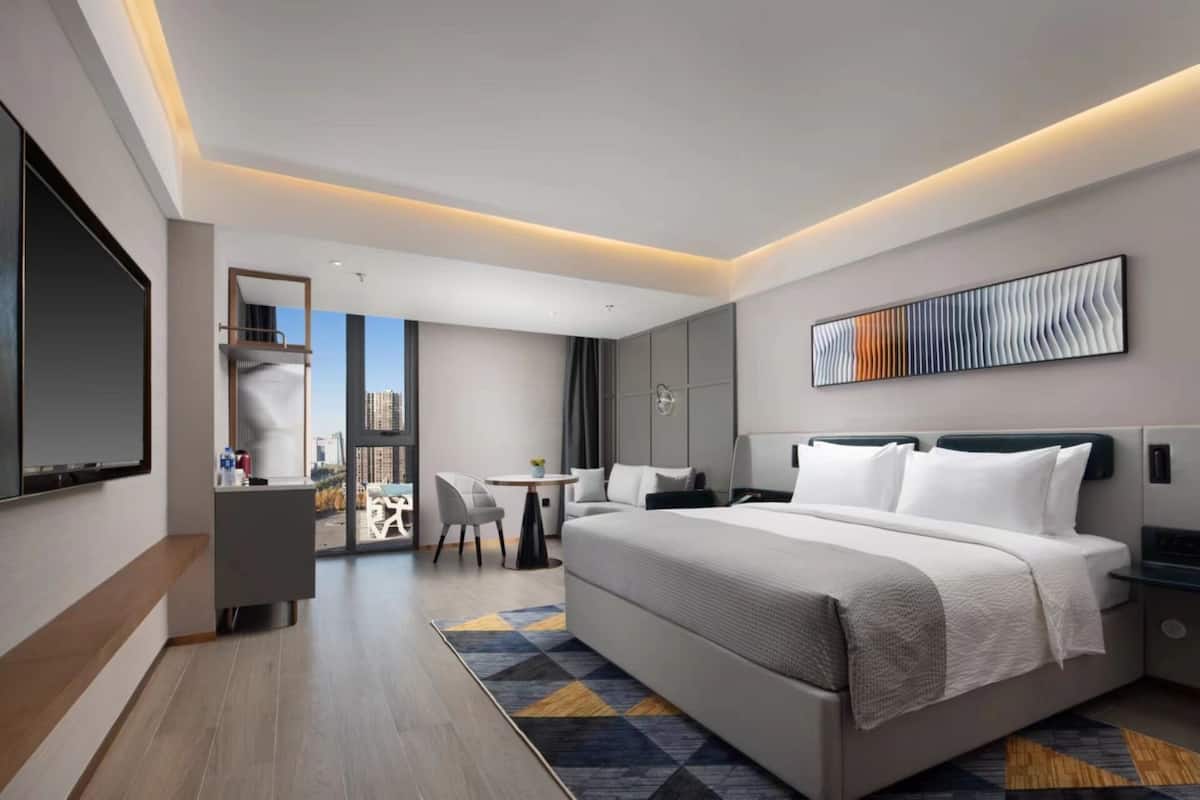 A modern hotel room with a large bed, a desk, a chair, and a view of the city.