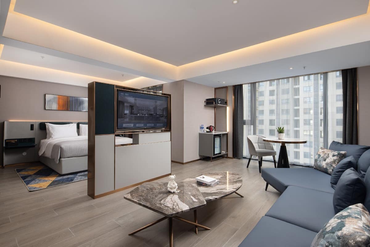 A modern hotel room with a bed, a TV, a desk, a sofa, and a large window.