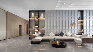 A modern lobby with a sectional sofa, a round coffee table, and built-in shelving with books and decorative items. - Wanda Yue Hotel Bengbu South Station (Bengbu)