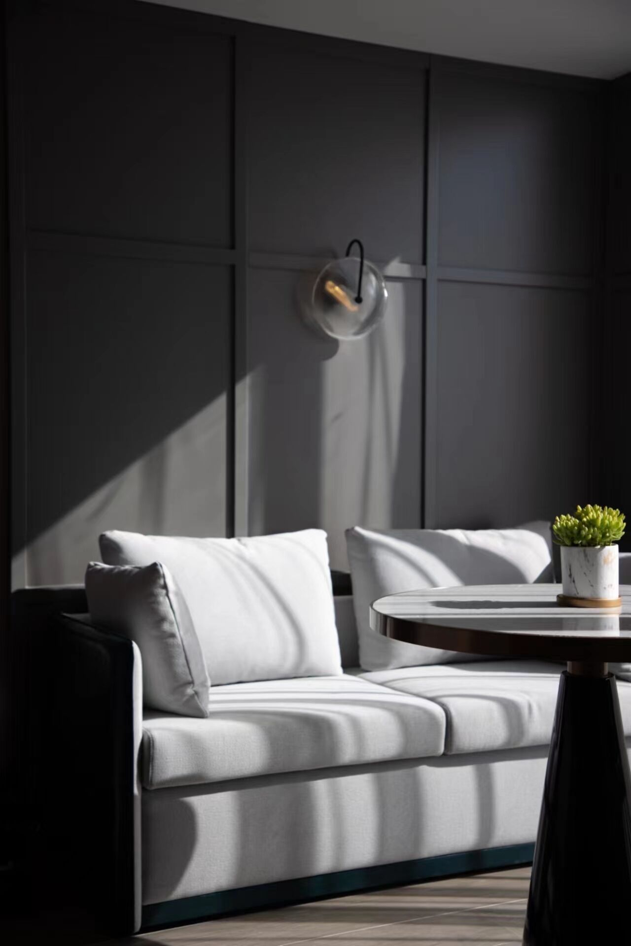 A modern, minimalist living room with a grey sofa, a small round table with a plant, and a wall-mounted light fixture.