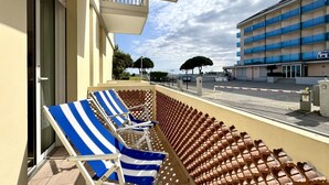 A balcony with striped lounge chairs and a tiled floor, overlooking a street with a hotel in the background.