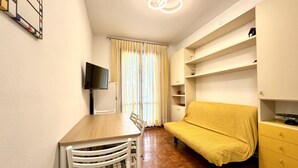 A compact living space with a wooden dining table, a sofa with a yellow cover, a TV mounted on the wall, and built-in shelving.