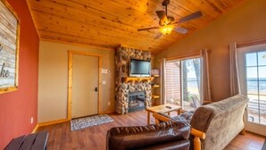 A cozy living room with a stone fireplace, wooden ceiling, and a large window offering a view of the beach. - Cabin 2 steps from the water (Isle)