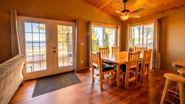 A wooden dining area with a table and chairs, large windows with curtains, and a ceiling fan. - Cabin 2 steps from the water (Isle)