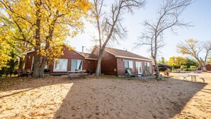 A cabin with a sandy ground, wooden exterior, and a large tree with yellow leaves. - Cabin 2 steps from the water (Isle)