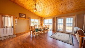 A spacious room with wooden floors, a dining area with chairs, a ceiling fan, and large windows offering a view of the outdoors. - 3 Bedroom Cabin 3 Beach front (Isle)