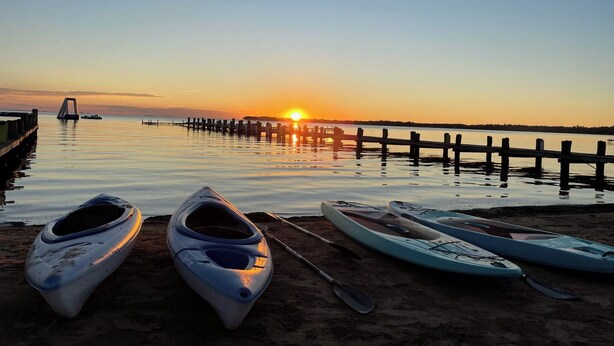 A beach with kayaks and a paddleboard, a pier extending into calm waters, and a sunset in the background. - Cabin 4 at the Lake - 2 bedrooms (Isle)