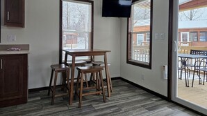 Una habitación con una mesa y sillas de madera, un televisor de pantalla plana en la pared y ventanas grandes con persianas.