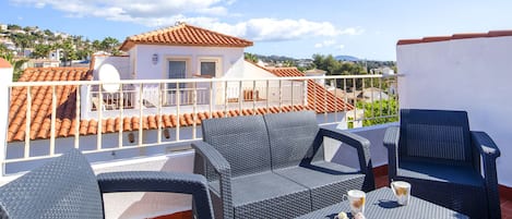 A rooftop terrace with wicker furniture, a table with cupcakes and a mug, and a view of a white building with orange tiles in the background.
