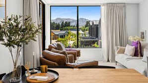 A modern living room with a large window, leather sofas, a wooden coffee table, and a view of mountains and buildings. - Mountainview Escape | Stylish, BBQ, and Parking (Wānaka)