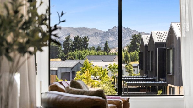 A room with a large window offering a view of a mountain range and residential buildings. - Mountainview Escape | Stylish, BBQ, and Parking (Wānaka)