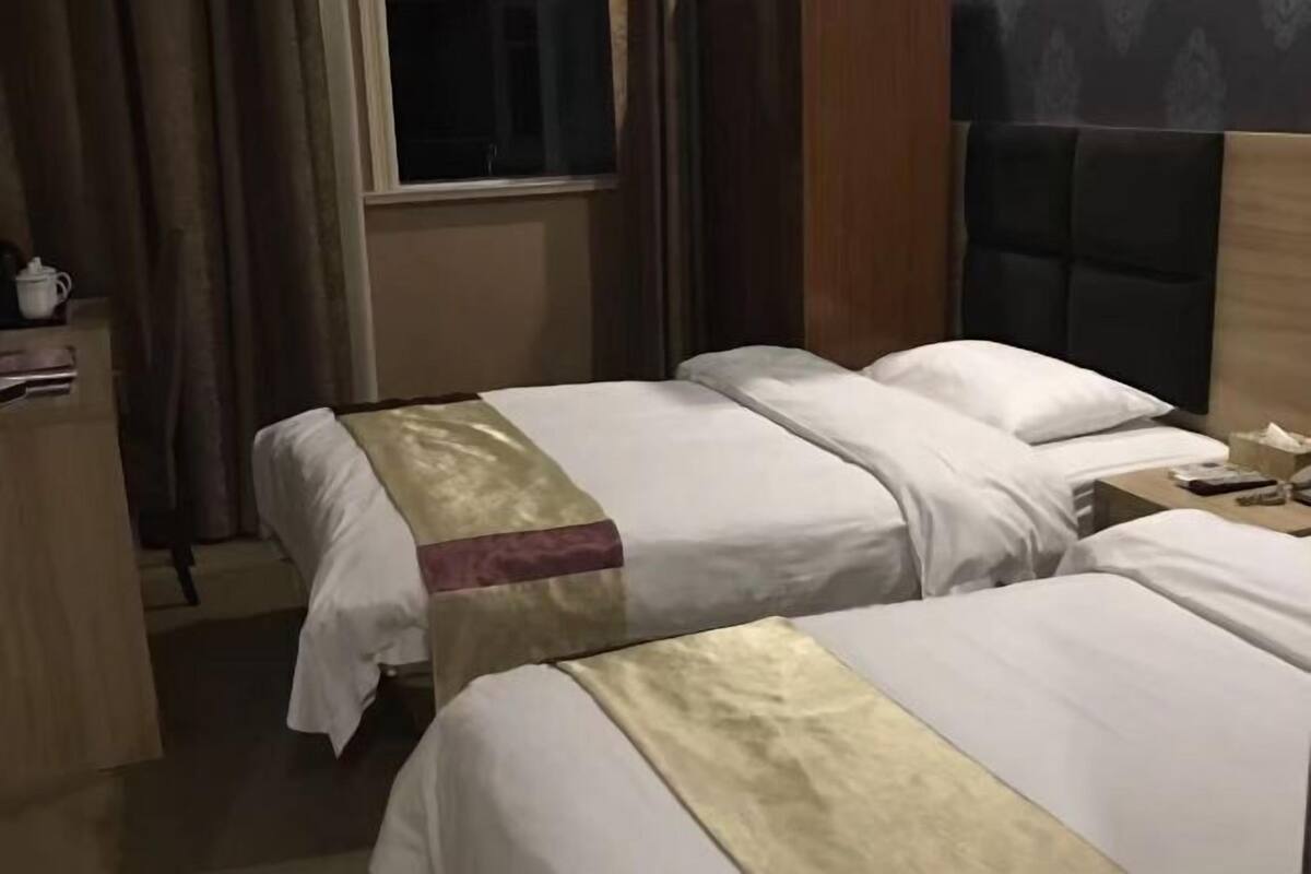 Ein Hotelzimmer mit zwei Betten, einem Holz-Kopfteil, einem Fenster mit Vorhängen und einem Nachttisch mit Fernbedienung.
