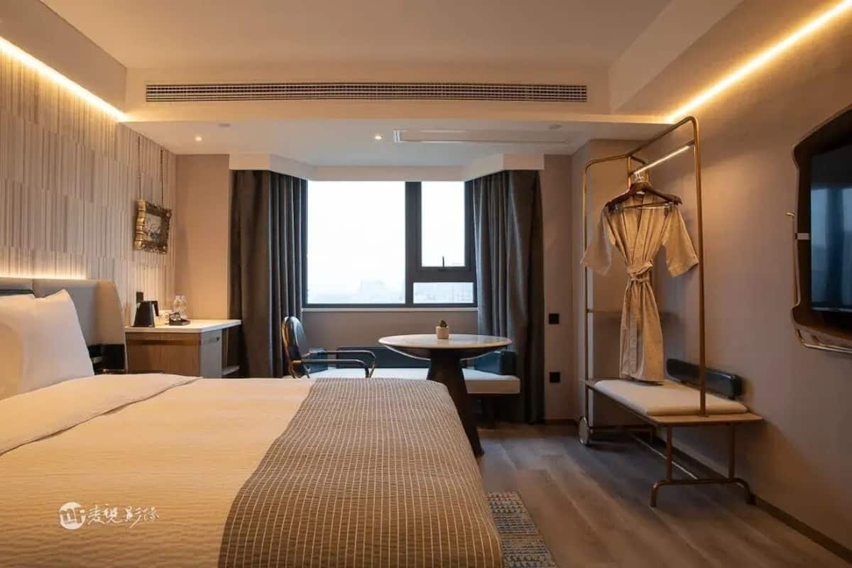 A hotel room with a large bed, a bedside table, a chair, a small round table, a hanging robe, and a large window.