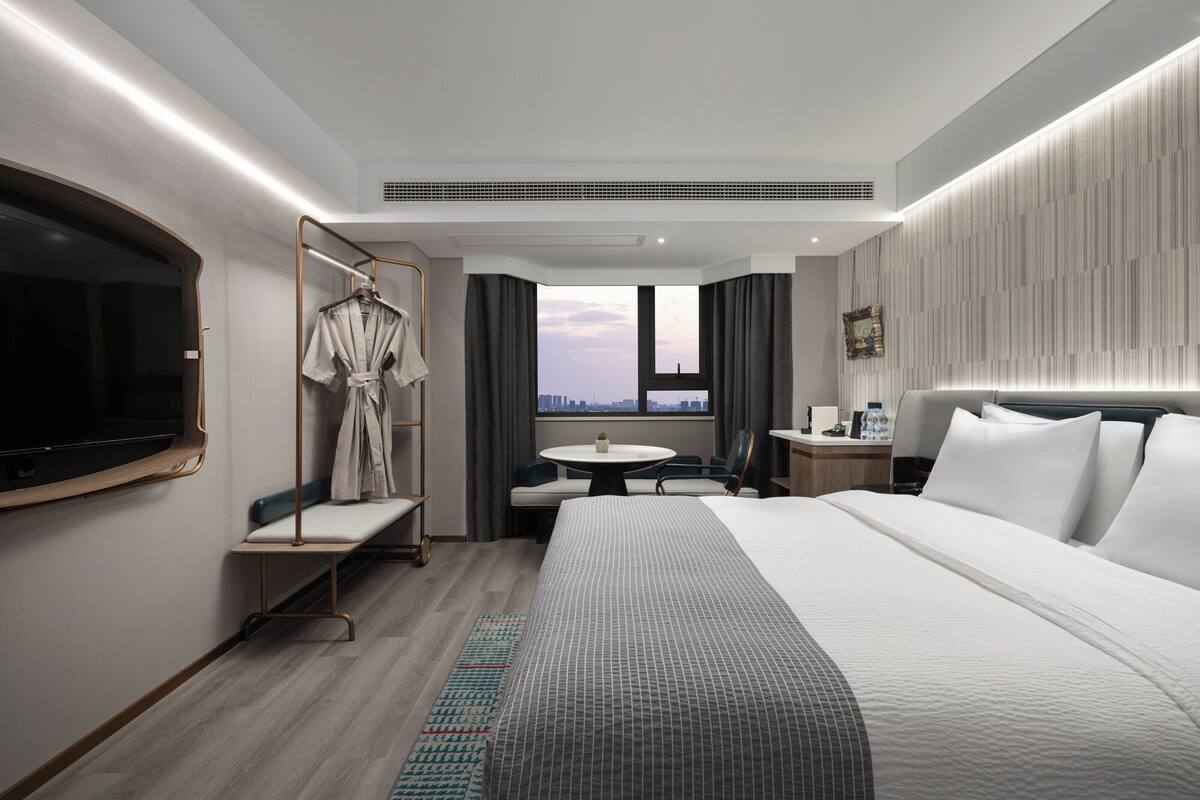A modern hotel room with a large bed, a TV, a seating area with a table, and a city view.